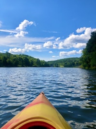 A vibrant kayak is floating on a calm, expansive body of water under a bright blue sky dotted with fluffy white clouds. Lush green trees line the horizon, creating a serene and picturesque natural landscape.
