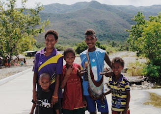 A group of five smiling children stands on a pathway in a rural setting, with lush green trees and hills in the background. One child is holding a large fish in a net. They are dressed in casual clothing and appear joyful and friendly.