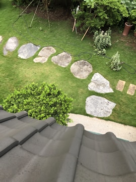 A view of a garden from an elevated position shows a series of large stepping stones arranged on a grassy lawn. Shrubs and small trees are planted around the garden, and there's evidence of gardening tools or ropes lying on the grass. In the foreground, a gray tiled roof is partially visible.