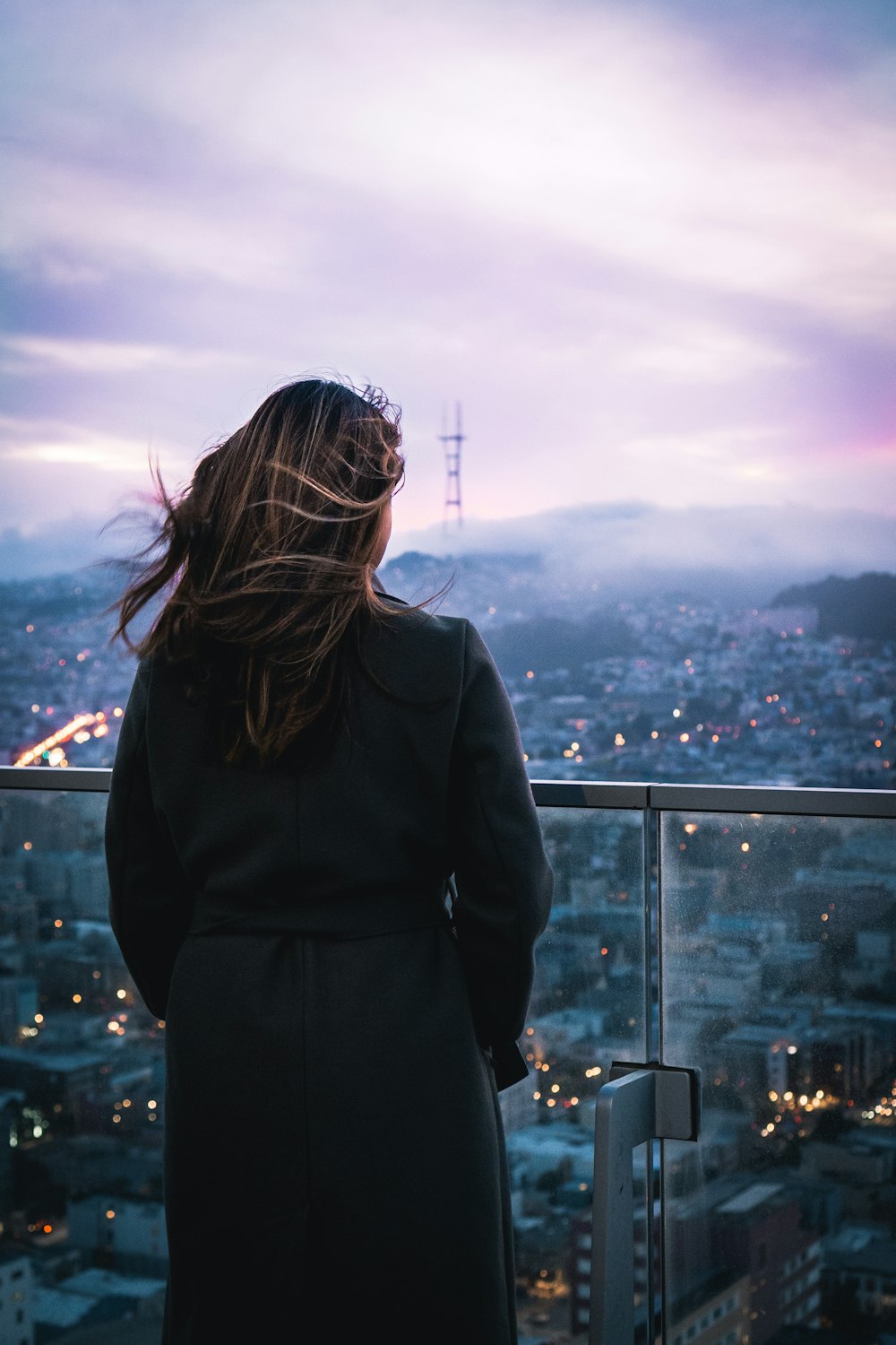 검은 코트를 입은 여자가 낮에 도시를 바라보는 건물 꼭대기에 서 있다
