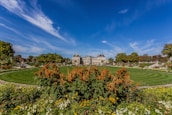 A well-maintained garden features a circular lawn surrounded by colorful flowers and neatly trimmed bushes in the foreground. In the background, a grand historic building with a slate roof stands majestically against a bright blue sky. Trees line the area, adding layers of green to the scenery.
