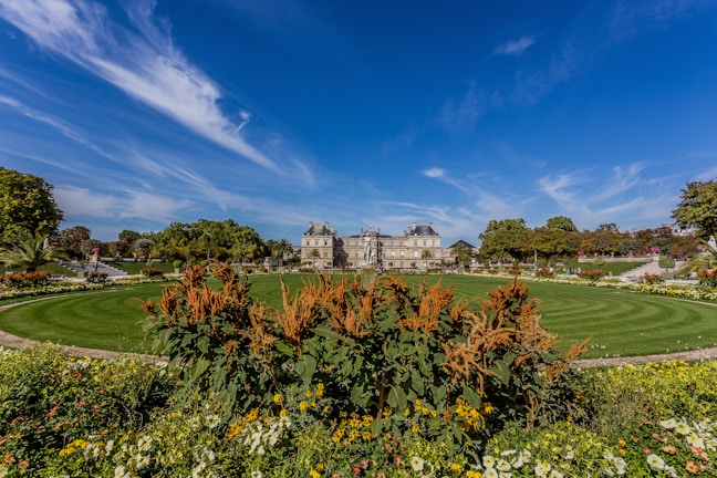 A well-maintained garden features a circular lawn surrounded by colorful flowers and neatly trimmed bushes in the foreground. In the background, a grand historic building with a slate roof stands majestically against a bright blue sky. Trees line the area, adding layers of green to the scenery.