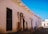 A rustic street scene with a row of white adobe buildings featuring wooden doors and ornamental ironwork. Several signs hang on the walls, including one for 'Artesanías'. Two people sit on the edge of the sidewalk, engaging in conversation. The environment is bright, with a clear blue sky overhead.