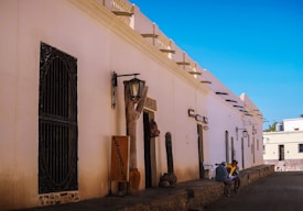 A rustic street scene with a row of white adobe buildings featuring wooden doors and ornamental ironwork. Several signs hang on the walls, including one for 'Artesan&iacute;as'. Two people sit on the edge of the sidewalk, engaging in conversation. The environment is bright, with a clear blue sky overhead.
