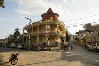 A round, two-story building with a red conical roof is prominently situated at a street intersection. Various posters are attached to its yellow exterior. Several people are walking nearby, and motorcycles along with a few parked vehicles, including a UN-marked car, can be seen. The surrounding area contains electrical poles with wires crossing the street.