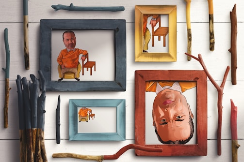Several artistic frames are arranged on a white wooden surface, each containing a colorful caricature of a man wearing an orange shirt, with one frame displaying the image upside down. Surrounding the frames, there are various sticks and branches of different colors and shapes.