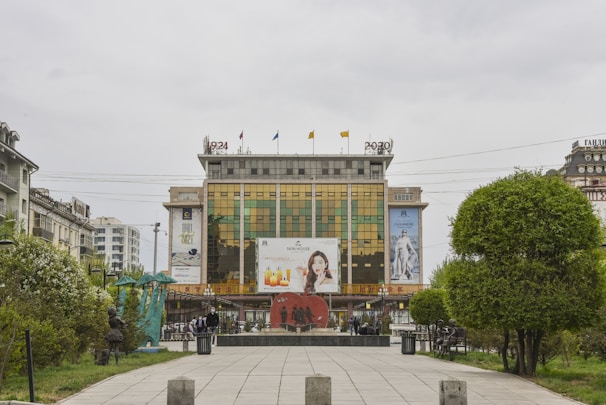 A multi-story commercial building with mirrored glass windows reflects its surroundings. The building features large advertising banners, including images promoting products and fashion. In front of the building is a walkway lined with trimmed trees and a statue. The sky is overcast, suggesting a cloudy day.
