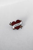 A pin shaped like a heart with a white banner that reads 'I LOVE ART'. The heart has brown arrows extending from its sides, and the background is a plain, light-colored surface.