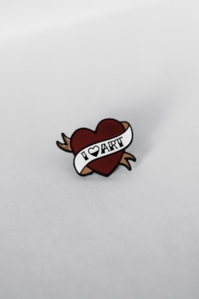 A pin shaped like a heart with a white banner that reads 'I LOVE ART'. The heart has brown arrows extending from its sides, and the background is a plain, light-colored surface.
