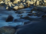 A stream flows over an assortment of smooth rocks, with water creating a soft, blurred effect as it moves. The boulders are rounded and varying in size, some submerged and others partly emerging above the water's surface. The lighting highlights the textures and shapes of the rocks, contrasting with the flowing water.
