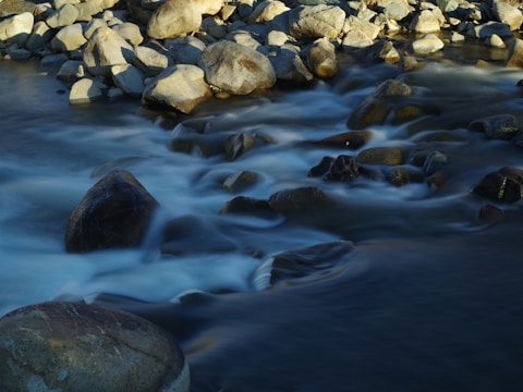 A stream flows over an assortment of smooth rocks, with water creating a soft, blurred effect as it moves. The boulders are rounded and varying in size, some submerged and others partly emerging above the water's surface. The lighting highlights the textures and shapes of the rocks, contrasting with the flowing water.