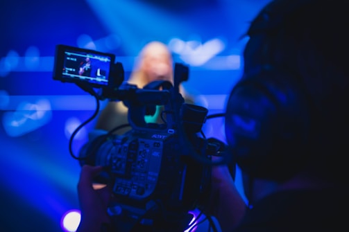 A person is operating a professional video camera with a display screen showing a close-up of a performer on stage. The scene is illuminated with a blue light, creating a vibrant and focused atmosphere. The camera is detailed, with visible buttons and a grip, and the background is blurred with blue hues.
