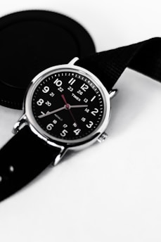 A black wristwatch with a round face featuring white numerals and red hour and minute hands. The watch is set against a light background, and a black camera lens cap is partially visible next to it. The strap of the watch is also black, adding to the monochromatic theme.