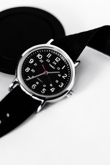 A black wristwatch with a round face featuring white numerals and red hour and minute hands. The watch is set against a light background, and a black camera lens cap is partially visible next to it. The strap of the watch is also black, adding to the monochromatic theme.