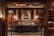A cozy and elegantly decorated room with wooden paneling and warm lighting. The space features a central sofa with colorful cushions, framed by two ornate lamps. A classic portrait hangs prominently on the wall behind the sofa, adding a touch of sophistication. Bookshelves line the right side, suggesting a library or study atmosphere.