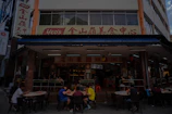 A street-side restaurant with several people seated at dark outdoor tables. The building has a sign written in both English and Chinese characters, with a prominent logo. Customers are casually dining and chatting, creating a lively atmosphere. The restaurant suggests an Asian culinary theme.