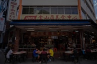 A street-side restaurant with several people seated at dark outdoor tables. The building has a sign written in both English and Chinese characters, with a prominent logo. Customers are casually dining and chatting, creating a lively atmosphere. The restaurant suggests an Asian culinary theme.