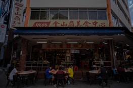 A street-side restaurant with several people seated at dark outdoor tables. The building has a sign written in both English and Chinese characters, with a prominent logo. Customers are casually dining and chatting, creating a lively atmosphere. The restaurant suggests an Asian culinary theme.