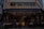 A street-side restaurant with several people seated at dark outdoor tables. The building has a sign written in both English and Chinese characters, with a prominent logo. Customers are casually dining and chatting, creating a lively atmosphere. The restaurant suggests an Asian culinary theme.