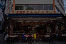 A street-side restaurant with several people seated at dark outdoor tables. The building has a sign written in both English and Chinese characters, with a prominent logo. Customers are casually dining and chatting, creating a lively atmosphere. The restaurant suggests an Asian culinary theme.