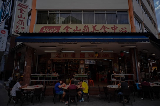 A street-side restaurant with several people seated at dark outdoor tables. The building has a sign written in both English and Chinese characters, with a prominent logo. Customers are casually dining and chatting, creating a lively atmosphere. The restaurant suggests an Asian culinary theme.