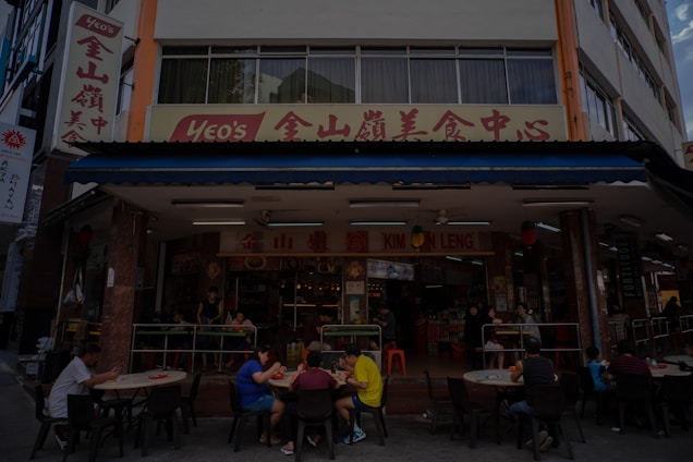 A street-side restaurant with several people seated at dark outdoor tables. The building has a sign written in both English and Chinese characters, with a prominent logo. Customers are casually dining and chatting, creating a lively atmosphere. The restaurant suggests an Asian culinary theme.
