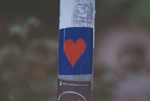 A red heart shape is painted on a blue rectangular background on a pole. The background is slightly weathered, and the pole has a textured appearance. There is a blurred background with green and brown tones, indicating foliage.