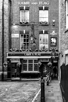 A historic brick building features the sign 'LAMB AND FLAG' above a public house entrance. The facade includes multiple paned windows adorned with small flower boxes. A traditional lantern hangs above the entrance, adding a vintage charm. The scene is monochrome, highlighting the texture and details of the brickwork and cobblestone street.
