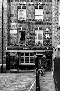 A historic brick building features the sign 'LAMB AND FLAG' above a public house entrance. The facade includes multiple paned windows adorned with small flower boxes. A traditional lantern hangs above the entrance, adding a vintage charm. The scene is monochrome, highlighting the texture and details of the brickwork and cobblestone street.