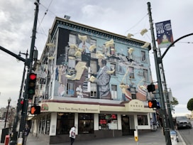 A vibrant mural covers the side of a building, depicting adults and children in various activities, surrounded by flying papers. The mural adorns the exterior of the New Sun Hong Kong Restaurant at a street corner. Traffic lights and street signs are visible, and people are walking by.