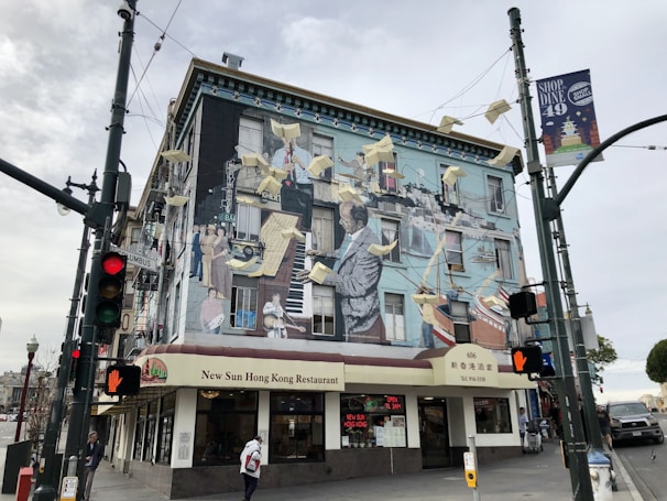 A vibrant mural covers the side of a building, depicting adults and children in various activities, surrounded by flying papers. The mural adorns the exterior of the New Sun Hong Kong Restaurant at a street corner. Traffic lights and street signs are visible, and people are walking by.