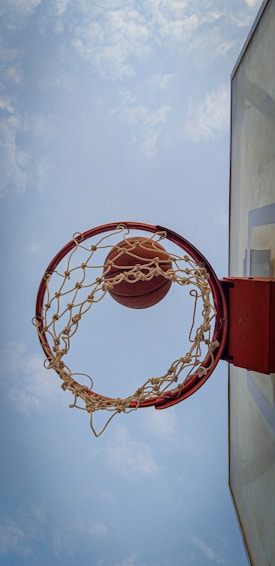 A basketball is mid-air heading towards the hoop from a bottom-up perspective. The net hangs down, creating a dynamic sense of motion. The background consists of a clear sky with a few clouds, emphasizing an outdoor setting.