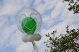 A balloon cluster with a transparent outer balloon decorated with white heart motifs surrounding a solid green balloon inside. Below, small white balloons form a base. They are positioned against a partly cloudy sky, with some tree branches visible in the background.