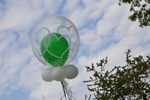 A balloon cluster with a transparent outer balloon decorated with white heart motifs surrounding a solid green balloon inside. Below, small white balloons form a base. They are positioned against a partly cloudy sky, with some tree branches visible in the background.