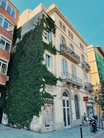 A multi-story building with traditional architecture features balconies with intricate railings. The left side is covered in lush green ivy, contrasting with the pale cream facade. A storefront with a sign reading 'Nails' and another marked 'FARMACIA' are visible at street level. A woman stands nearby with a bicycle and a motorcycle parked in front.