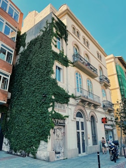 A multi-story building with traditional architecture features balconies with intricate railings. The left side is covered in lush green ivy, contrasting with the pale cream facade. A storefront with a sign reading 'Nails' and another marked 'FARMACIA' are visible at street level. A woman stands nearby with a bicycle and a motorcycle parked in front.
