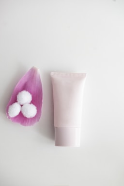 A minimalist arrangement featuring a soft pink flower petal holding three small white cotton balls on the left and a neutral-toned, cylindrical skincare product tube on the right. The background is a smooth white surface, enhancing the clean and calm composition.