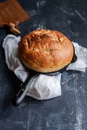 A freshly baked round loaf of bread sits in a cast iron skillet, which is placed on top of a white cloth. The background has a dark, rustic wooden texture, and there is a wooden cutting board partially visible in the upper left corner.