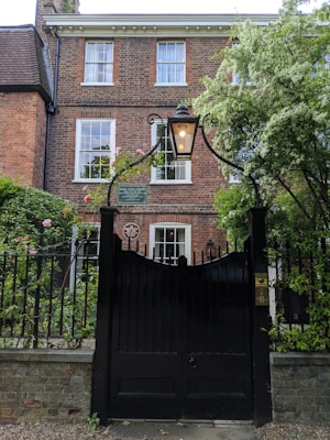 A historic brick house with white-trimmed windows, surrounded by lush greenery, is visible behind a decorative black iron gate with a lantern on top. The garden features blooming flowers and trees, contributing to a serene atmosphere.