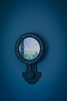 A vintage wall clock with Roman numerals and a pendulum style design is mounted against a plain wall. The clock face is surrounded by ornate detailing and a dark finish.