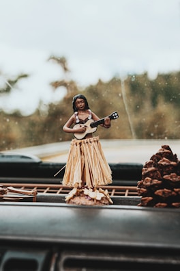 A dashboard with a small hula dancer figurine featuring a straw skirt and a ukulele. The background is a blurred outdoor view and a large pinecone is visible next to the figurine.
