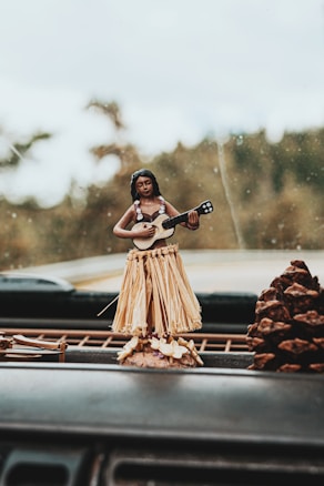 A dashboard with a small hula dancer figurine featuring a straw skirt and a ukulele. The background is a blurred outdoor view and a large pinecone is visible next to the figurine.