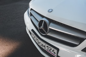 A close-up view of the front grille and logo of a white Mercedes-Benz vehicle. The license plate is visible, featuring black text on a white background.