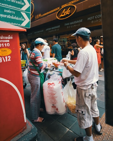 A street vendor and a customer are engaged in a transaction in a bustling urban area. The vendor is preparing food, surrounded by a variety of supplies, including large bags. Behind them are signs in various languages and people walking by. The setting is lively and busy.