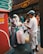 A street vendor and a customer are engaged in a transaction in a bustling urban area. The vendor is preparing food, surrounded by a variety of supplies, including large bags. Behind them are signs in various languages and people walking by. The setting is lively and busy.