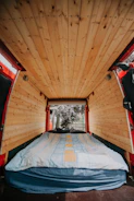 A cozy van interior with wooden paneling on the ceiling and walls. A bed with a blue and yellow striped blanket is neatly arranged at the back, leading to an open rear door where trees and a fence are visible outside.