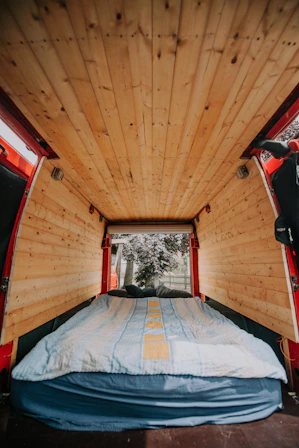 A cozy van interior with wooden paneling on the ceiling and walls. A bed with a blue and yellow striped blanket is neatly arranged at the back, leading to an open rear door where trees and a fence are visible outside.