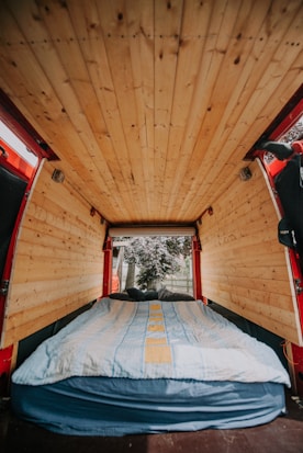 A cozy van interior with wooden paneling on the ceiling and walls. A bed with a blue and yellow striped blanket is neatly arranged at the back, leading to an open rear door where trees and a fence are visible outside.
