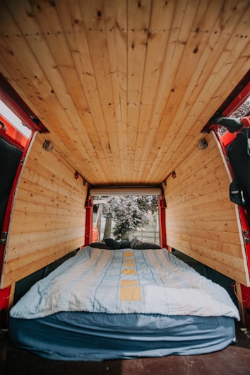 A cozy van interior with wooden paneling on the ceiling and walls. A bed with a blue and yellow striped blanket is neatly arranged at the back, leading to an open rear door where trees and a fence are visible outside.