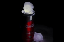 A can of Coca-Cola Plus Coffee rests on a dark surface. An ice cube is placed on top of the can's open tab, while another ice cube lies beside the can. The dim lighting creates a dramatic contrast, highlighting the red and steel grey colors of the can.